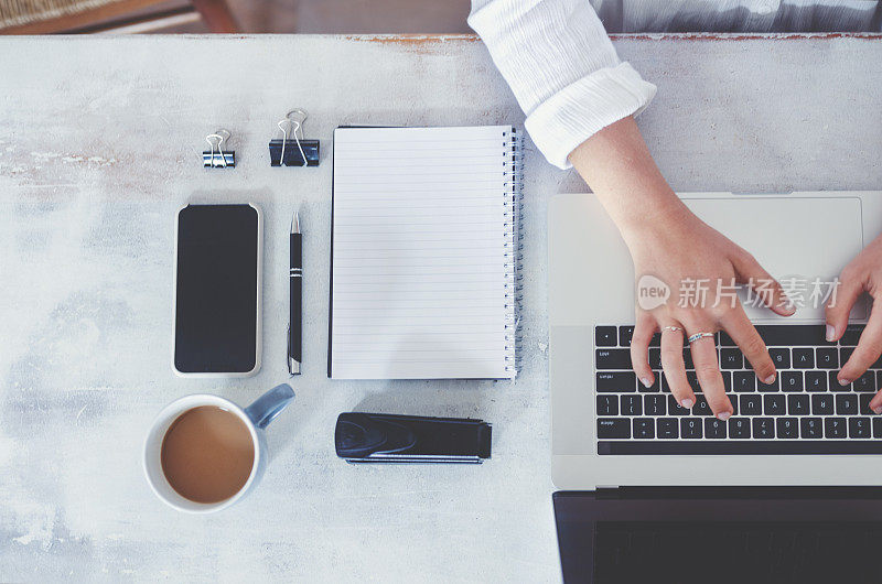 Top view of woman using laptop while sitting at the table with laptop, mobile phone, blank note pad and pen.女人使用笔记本电脑时的俯视图。